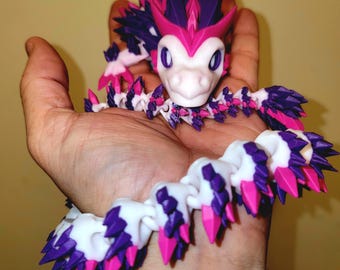 May include: A pink and purple dragon-shaped bracelet made of plastic. The bracelet is made up of small, interlocking pieces that form a chain. The dragon's head is at the top of the bracelet, and its tail is at the bottom.