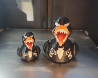 May include: Two black rubber duck toys with white eyes and teeth, and an orange beak and tongue. The ducks have a white spider-web design on their chest. One duck is smaller than the other.