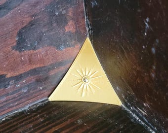 May include: A gold-colored triangular corner bracket featuring a central sunburst design with radiating lines and a small circular indentation in the middle, positioned to fit into the interior corner of a dark brown wooden structure.