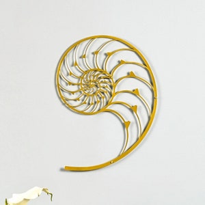 Peut inclure: Une sculpture murale en métal doré en forme de coquille de nautile. La sculpture est accrochée à un mur blanc au-dessus d'une table en bois avec un vase de lys blancs et un livre.