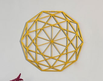 May include: A yellow geometric wall decoration with a diamond shape design. The decoration is hanging on a light grey wall above a wooden table with a vase of purple flowers and a book.