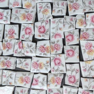 May include: A collection of white ceramic tile shards featuring a floral pattern with pink and yellow roses and green leaves. Each piece is irregularly shaped with smooth edges, displaying various sections of the repeating botanical design.