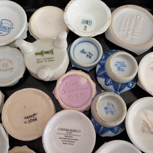 May include: A collection of small, white ceramic dishes and saucers, some with blue or gold accents and brand names like Gibson, Corelle, and Cynthia Rowley. Some pieces are stamped with "Made in Japan" or "Made in China".