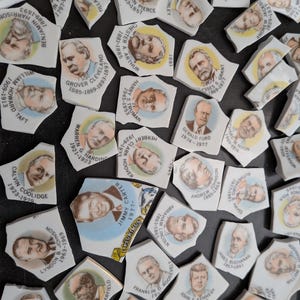 May include: A collection of white ceramic shards featuring color portraits of United States Presidents. Each piece displays a name, term years, and a bust illustration. One fragment includes the text E PLURIBUS UNUM. The shards vary in shape and size.