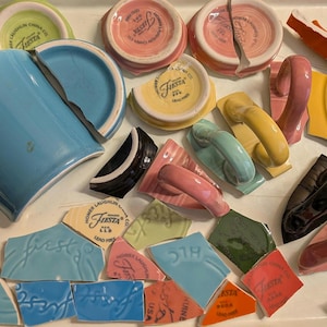 May include: A collection of broken Fiesta ware pottery pieces in various colors, including blue, pink, yellow, and green. The fragments include mug handles, plates, and cup bases, some with the "Fiesta" logo. The pieces are arranged on a white surface.