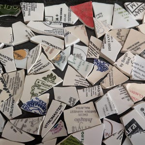 May include: A collection of broken ceramic pieces, each with fragments of text and designs. The shards vary in size and shape, with a mix of white, cream, and red hues. Some pieces display floral patterns and brand names.