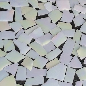 May include: An overhead shot displays numerous irregularly shaped, iridescent glass pieces scattered across a dark surface. The glass fragments vary in size and shape, with a mix of white, pale yellow, and light blue hues.