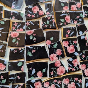 May include: A collection of broken ceramic tile pieces featuring a black background with a floral pattern of pink roses, small pink flowers, and light blue leaves. Each individual shard has a thin gold trim along the curved edge of the ceramic.