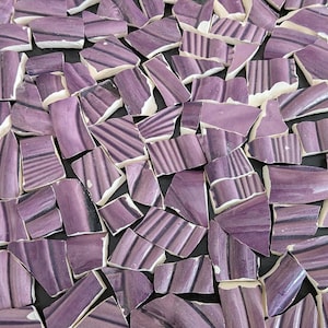 May include: A collection of broken ceramic tiles in shades of purple and white. Each piece has a unique shape and features a striped pattern in darker purple hues. The tiles are scattered across a dark surface.