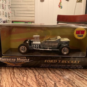 May include: A die-cast metal Ford T-Bucket model car in a display box. The model is silver and black with chrome details. The box has the text "American Muscle" and "Ford T-Bucket". A "NEW Tool!" sticker is on the box.
