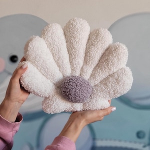 May include: A white plush seashell cushion with a purple centre. The cushion is shaped like a seashell with a ruffled edge.