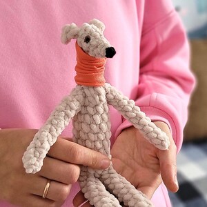 May include: A handmade, light gray crocheted dog toy with an orange neck warmer and a black nose. The dog has long, slender limbs and is held in someone's hands. The toy is made of a soft, textured yarn, and the overall aesthetic is cute and whimsical.