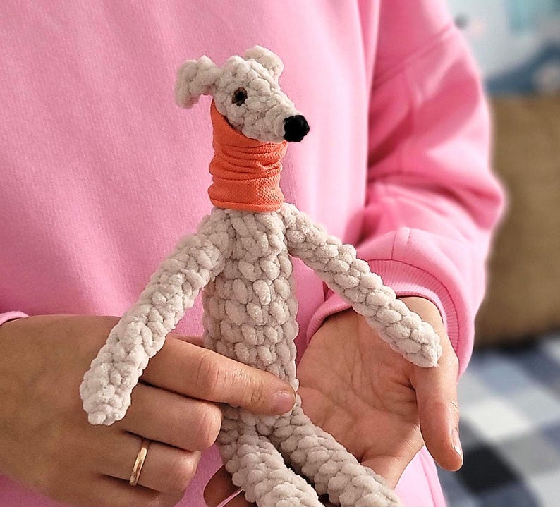 May include: A handmade, light gray crocheted dog toy with an orange neck warmer and a black nose. The dog has long, slender limbs and is held in someone's hands. The toy is made of a soft, textured yarn, and the overall aesthetic is cute and whimsical.