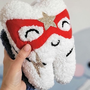 May include: White plush tooth with a red and gold superhero mask. The tooth has black eyes and a gold lightning bolt on the side.