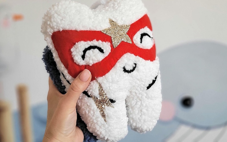 May include: White plush tooth with a red and gold superhero mask. The tooth has black eyes and a gold lightning bolt on the side.