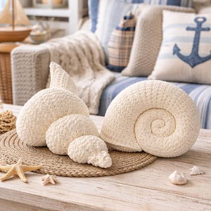May include: Cream-coloured, seashell-shaped decorative cushions on a woven mat. The cushions vary in size and shape, with a large spiral shell and smaller, textured forms. The scene is set on a light wood table with starfish and seashells, evoking a coastal theme.