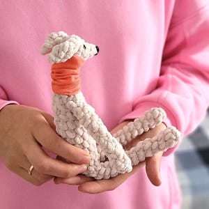 May include: A handmade, plush dog toy in cream-colored yarn with an orange neck wrap. The dog has a black nose and is held in a person's hands. The background is a soft pink color.