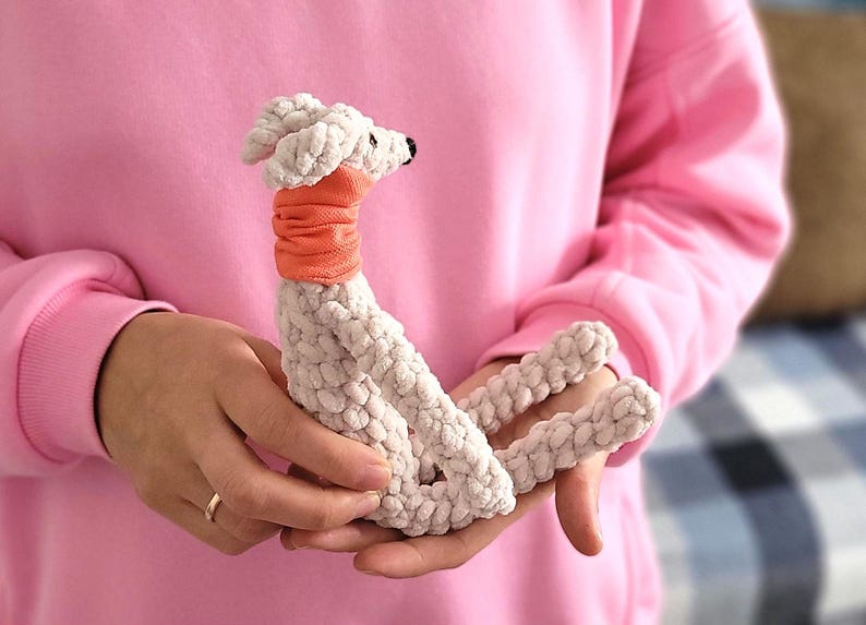 May include: A handmade, plush dog toy in cream-colored yarn with an orange neck wrap. The dog has a black nose and is held in a person's hands. The background is a soft pink color.