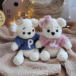 Può includere: Due orsacchiotti di peluche bianchi all'uncinetto che indossano maglioni. L'orsacchiotto a sinistra indossa un maglione blu con una lettera bianca "P" sopra. L'orsacchiotto a destra indossa un maglione rosa con una lettera bianca "D" sopra.