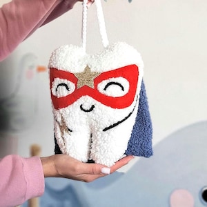 May include: A white plush tooth with a red and gold superhero mask and a blue cape. The tooth has a smiling face and a gold star on its mask.