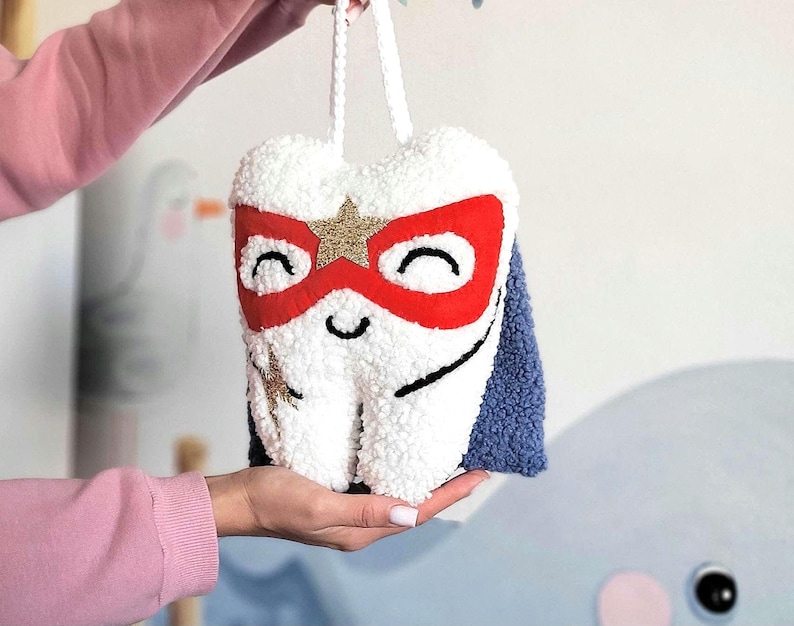 May include: A white plush tooth with a red and gold superhero mask and a blue cape. The tooth has a smiling face and a gold star on its mask.