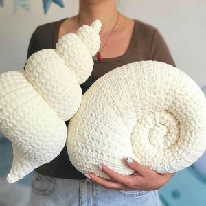 Op de afbeelding: Een groot, crèmekleurig, gehaakt schelpvormig kussen. Het kussen heeft een spiraalvormig ontwerp en een getextureerd oppervlak. Het kussen is zacht en pluche, geschikt voor woondecoratie.
