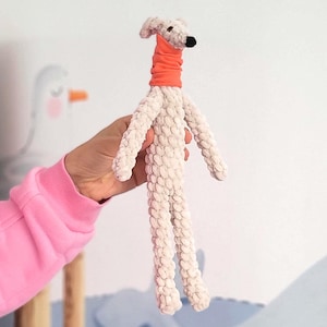 May include: A handmade plush dog toy in cream-colored fabric with long legs and arms. The dog has a small head with a black nose and is wearing an orange neckerchief. The toy is held in a person's hand.