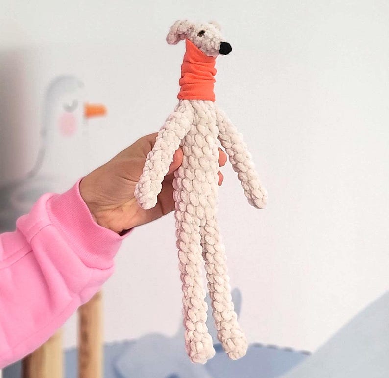 May include: A handmade plush dog toy in cream-colored fabric with long legs and arms. The dog has a small head with a black nose and is wearing an orange neckerchief. The toy is held in a person's hand.
