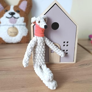 May include: A handmade white stuffed dog with a coral scarf, positioned in front of a wooden house-shaped decoration with a lavender roof. A corgi-shaped felt decoration is in the background.