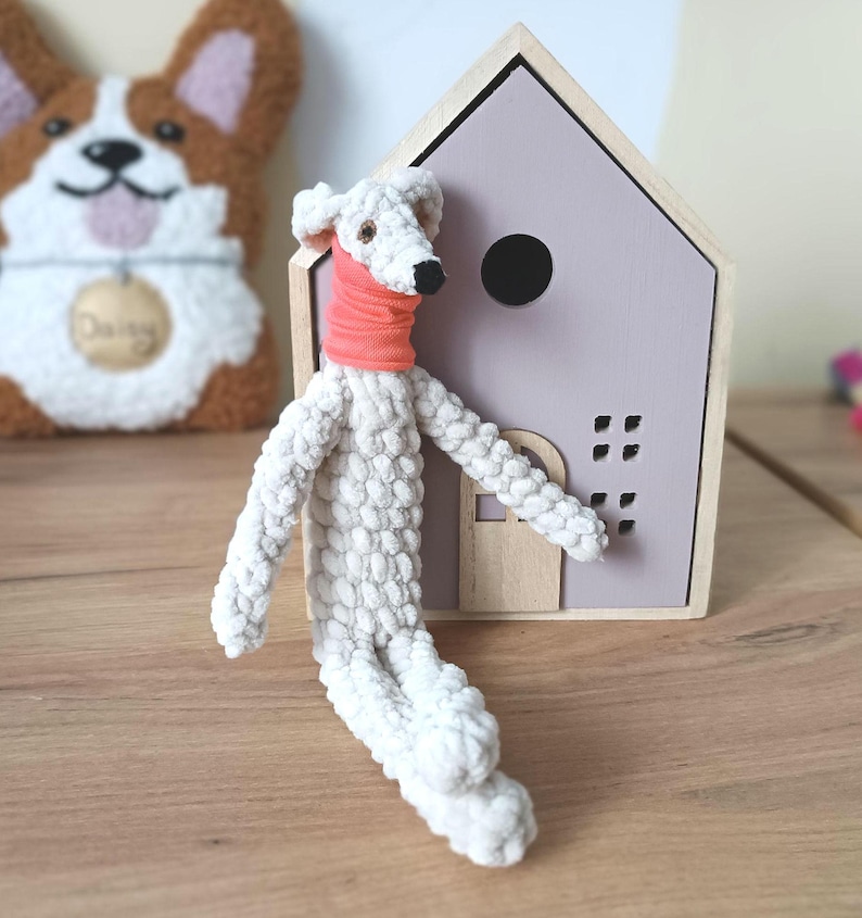 May include: A handmade white stuffed dog with a coral scarf, positioned in front of a wooden house-shaped decoration with a lavender roof. A corgi-shaped felt decoration is in the background.