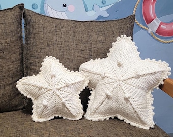 Puede incluir: Dos cojines blancos tejidos en forma de estrella de mar de diferentes tamaños descansan sobre un sofá marrón. Los cojines tienen un diseño texturizado y detallado. El fondo incluye un mural de temática náutica.