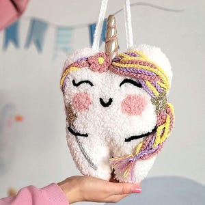May include: A white plush tooth shaped like a unicorn with a gold horn, pink cheeks, and purple and yellow braided hair.