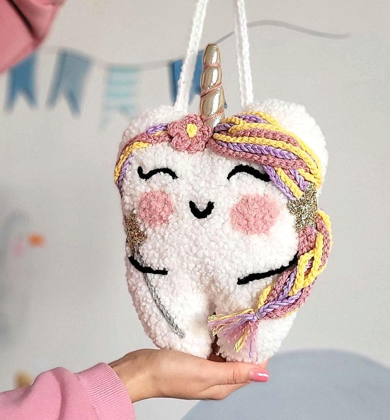 May include: A white plush tooth shaped like a unicorn with a gold horn, pink cheeks, and purple and yellow braided hair.