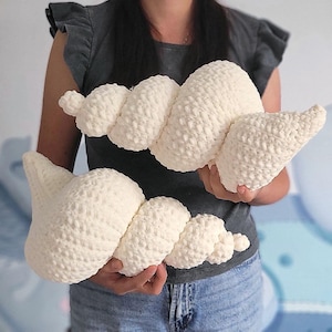 Puede incluir: Dos almohadas en forma de concha, de color blanco roto y hechas a ganchillo. Las almohadas tienen una superficie texturizada y con bultos y un diseño en espiral. Las almohadas están sujetas por una persona.