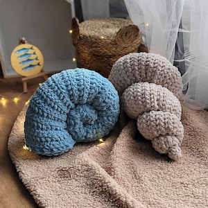 以下が含まれることがあります： 貝殻の形をしたニットのぬいぐるみ枕が2つ。青い貝殻の枕が左に、ベージュの貝殻の枕が右にあります。枕は薄い茶色のふわふわの毛布の上に置かれています。