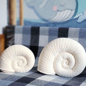 Puede incluir: Dos cojines decorativos en forma de concha, de color blanquecino. Los cojines tienen un diseño en espiral y una superficie texturizada. Se colocan sobre una manta a cuadros azules y blancos. El fondo presenta una ilustración de ballena.