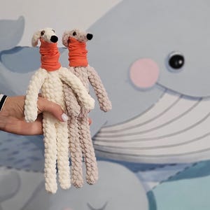 May include: Two handmade, stuffed dog dolls. One is cream-colored, the other is light brown. Both have orange necks and white heads with black eyes. The dolls are held in front of a wall mural featuring a blue whale and other aquatic elements.