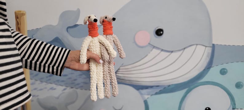 May include: Two handmade, stuffed dog dolls. One is cream-colored, the other is light brown. Both have orange necks and white heads with black eyes. The dolls are held in front of a wall mural featuring a blue whale and other aquatic elements.