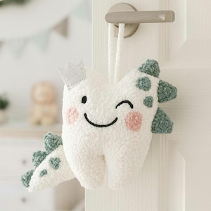 May include: A plush, white tooth-shaped door hanger with a silver crown, winking eye, and pink cheeks. The tooth has green spotted accents and is hanging from a door handle. A cute and whimsical design for a child's room.