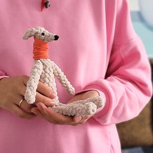 May include: A handmade, light gray crocheted dog with an orange neck warmer. The dog has a black nose and is held in someone's hands. The background is a pink sweatshirt.