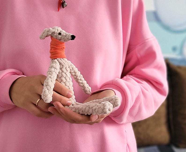May include: A handmade, light gray crocheted dog with an orange neck warmer. The dog has a black nose and is held in someone's hands. The background is a pink sweatshirt.