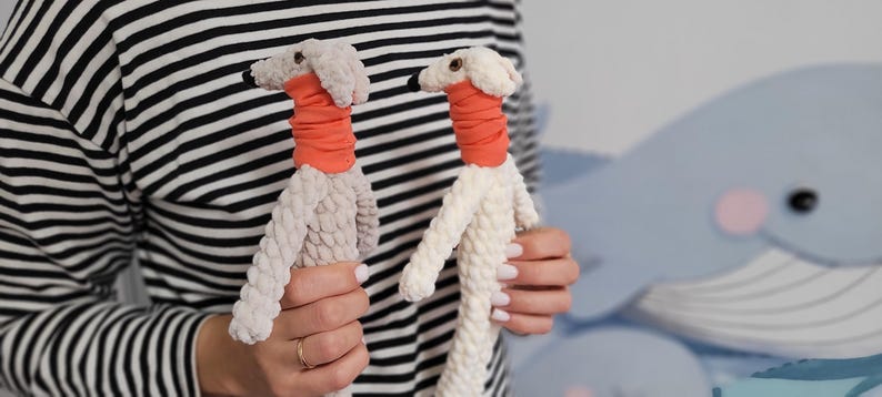 May include: Two plush dog toys. One is light gray, the other is white. Both have orange neckbands and black eyes. The toys are held up against a background with a whale design.