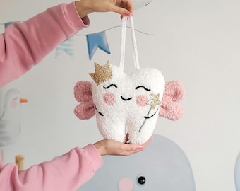 May include: A white plush tooth with pink wings and a gold crown. The tooth has a smiling face and a gold star on its cheek. The tooth is hanging from a white string.