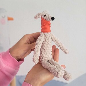 May include: A handmade plush dog toy in light gray with an orange neck warmer. The dog has a black nose and small, floppy ears. The toy is made of a soft, textured material and is being held by a person.