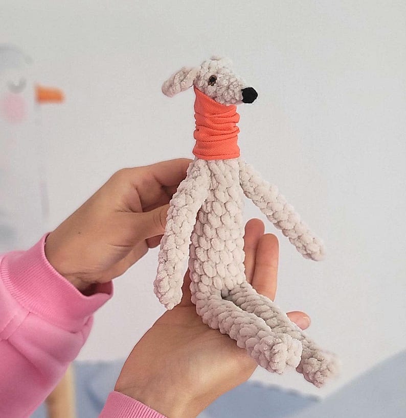 May include: A handmade plush dog toy in light gray with an orange neck warmer. The dog has a black nose and small, floppy ears. The toy is made of a soft, textured material and is being held by a person.