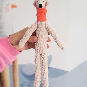 May include: A handmade plush dog toy with a cream-colored body and an orange neck warmer. The dog has a black nose and small, floppy ears. The toy is held by a person wearing a pink long-sleeved shirt.