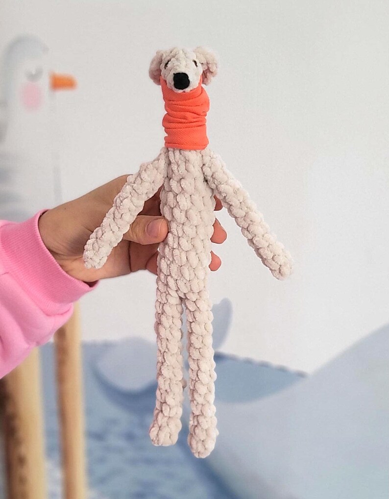 May include: A handmade plush dog toy with a cream-colored body and an orange neck warmer. The dog has a black nose and small, floppy ears. The toy is held by a person wearing a pink long-sleeved shirt.
