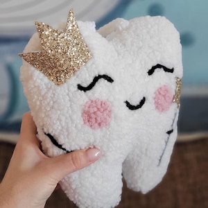 May include: A white plush tooth with a pink and black smiling face, wearing a gold glitter crown.