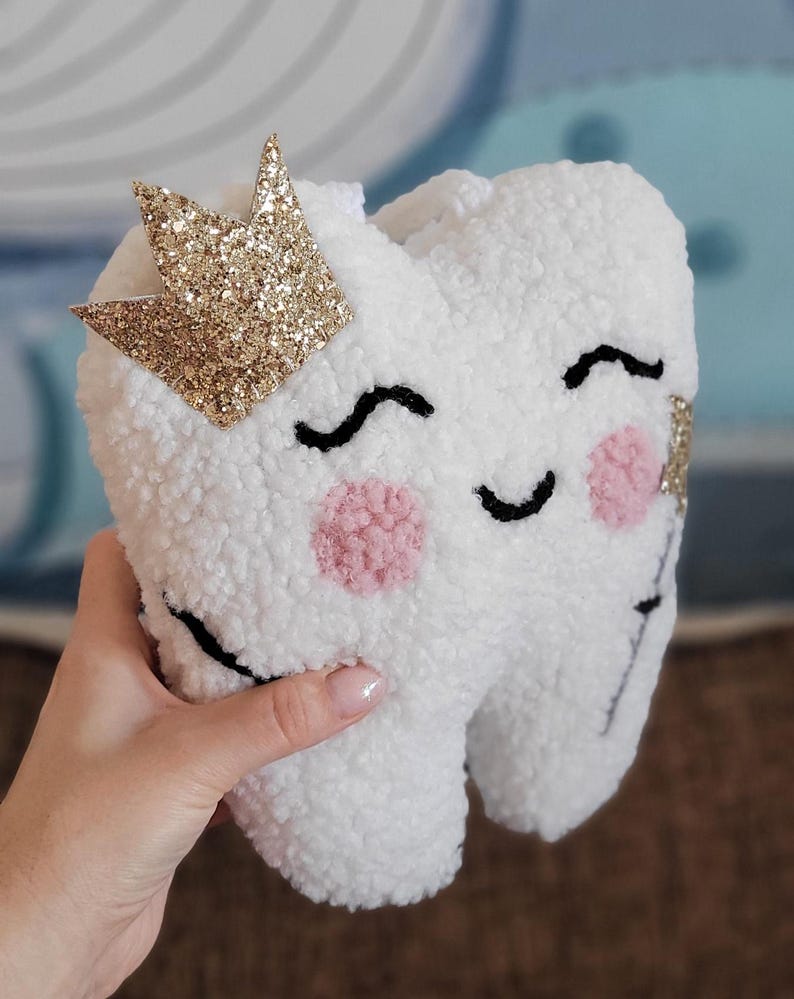 May include: A white plush tooth with a pink and black smiling face, wearing a gold glitter crown.