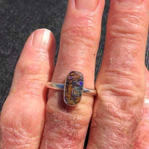 May include: A silver ring with an oval gemstone, displaying a pattern of brown, gold, purple, and blue. The ring is worn on a finger, highlighting the stone's intricate details. The ring's design is eye-catching.